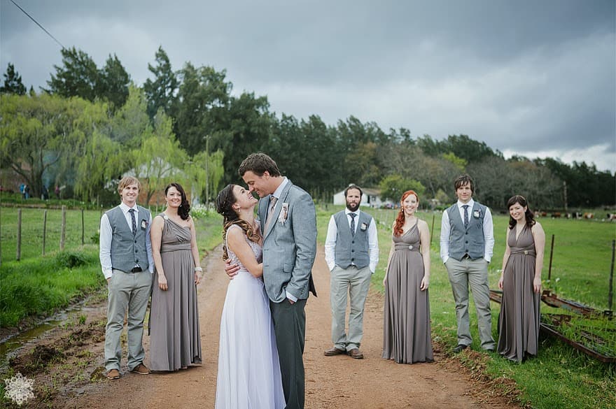 A bridal party on a farm road at Muldersvlei, the bride and groom kissing at the centre, willow trees and green paddocks behind