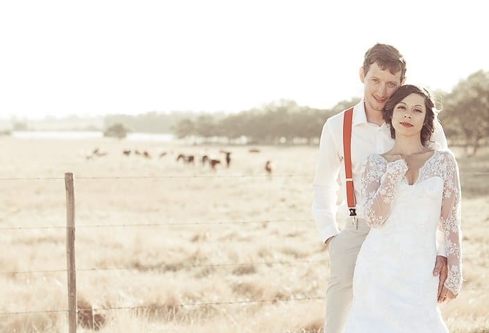 A newly married couple standing in a golden field on the farm, cattle grazing beyond the fence
