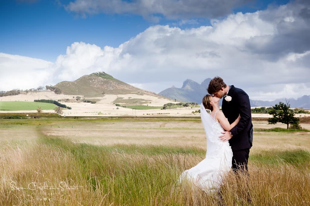 A bride and groom kissing in the open grasslands at Muldersvlei, mountains and a big Cape sky behind them