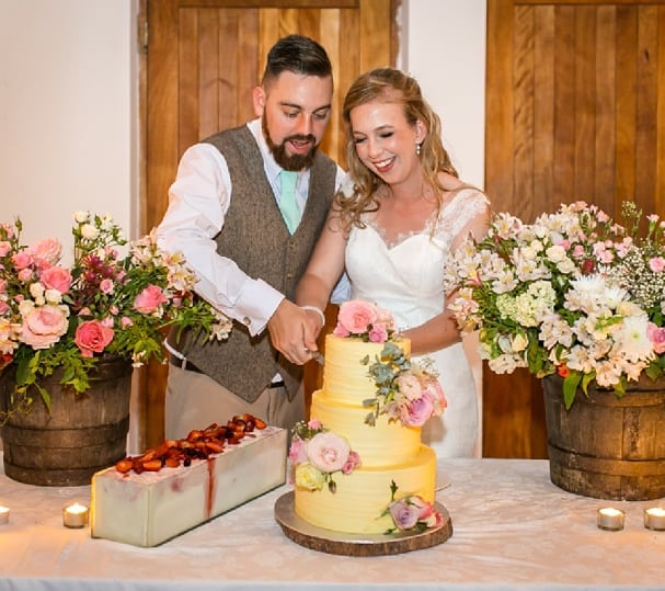 A bride and groom cutting their wedding cake at the reception at Muldersvlei, flowers in wine barrels behind them