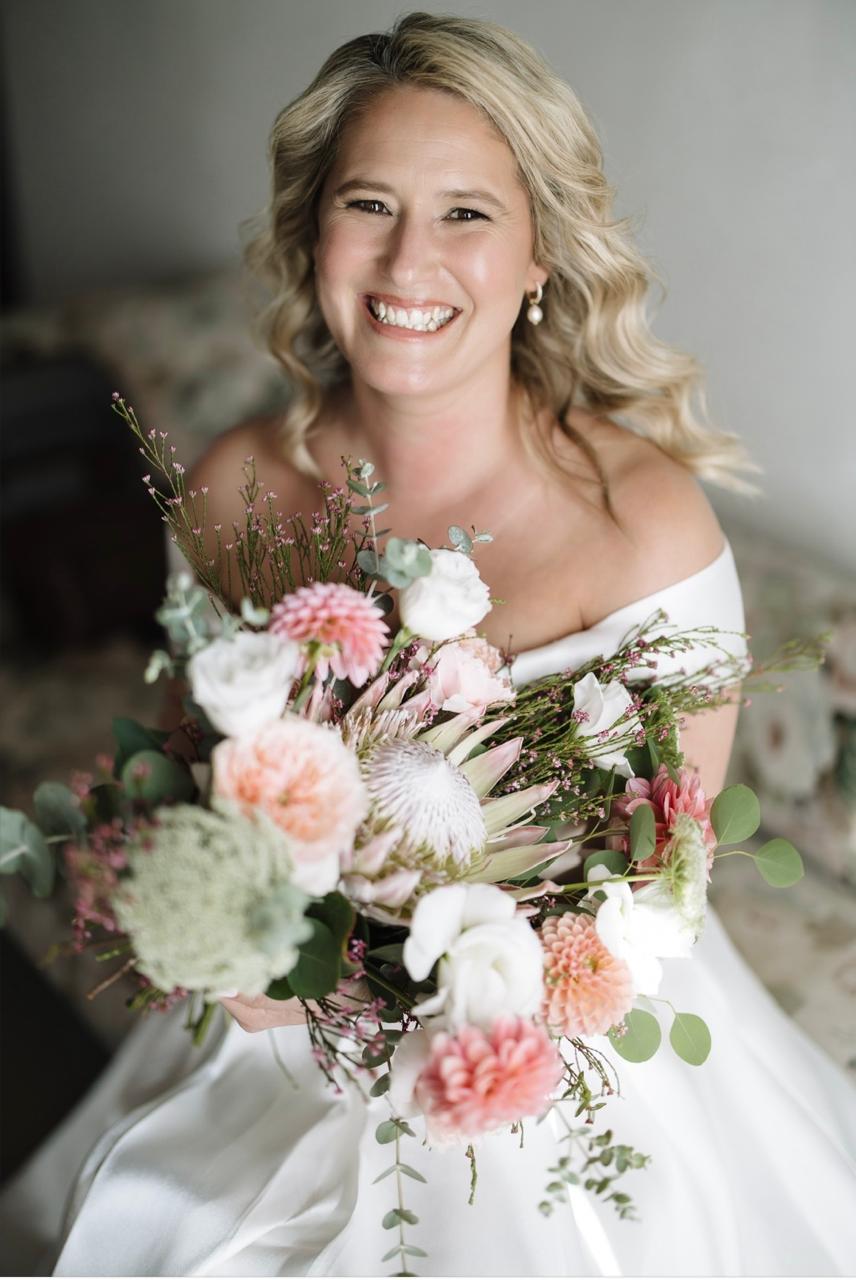 A bride smiling over her bouquet of king protea, dahlias and garden roses at Muldersvlei